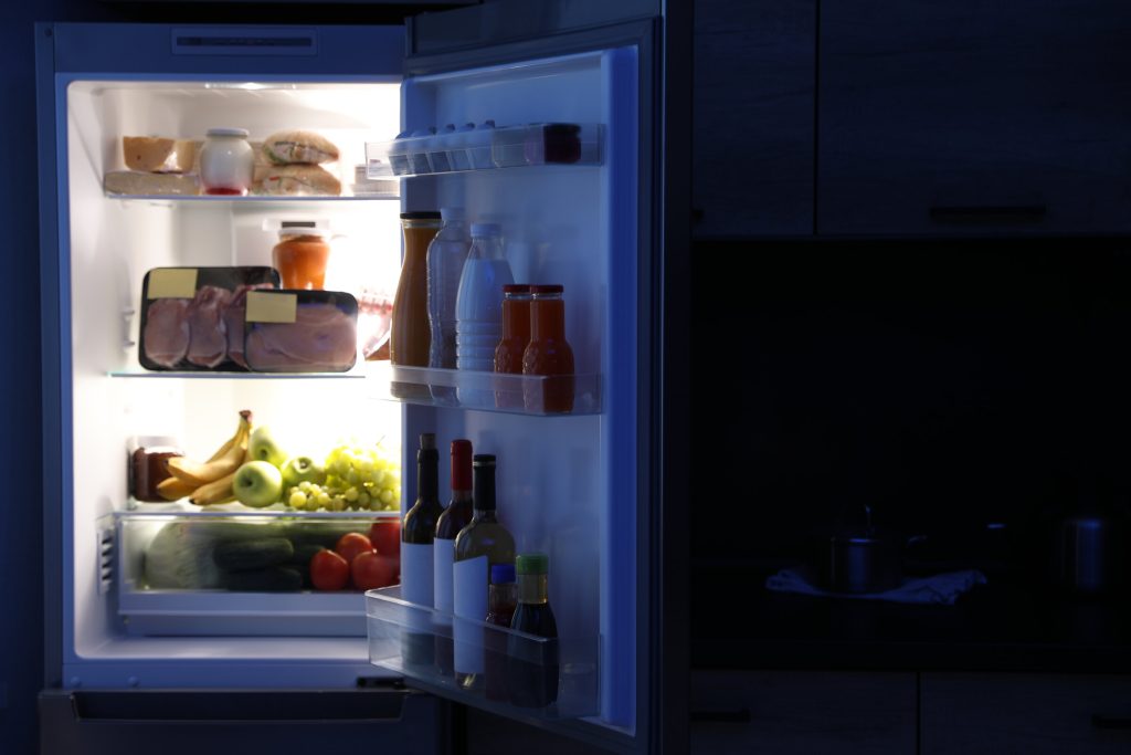 An open refrigerator lighting a dark kitchen at night, its shelves stocked with food