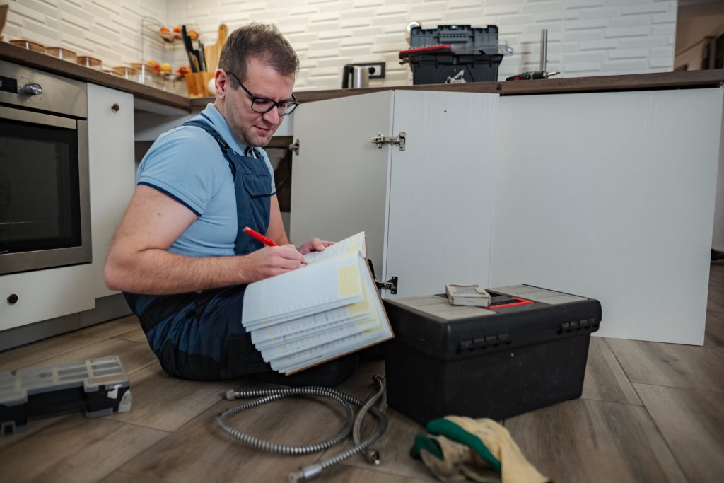 A technician sitting on a kitchen floor writing up a job on a clipboard beside an open tool box