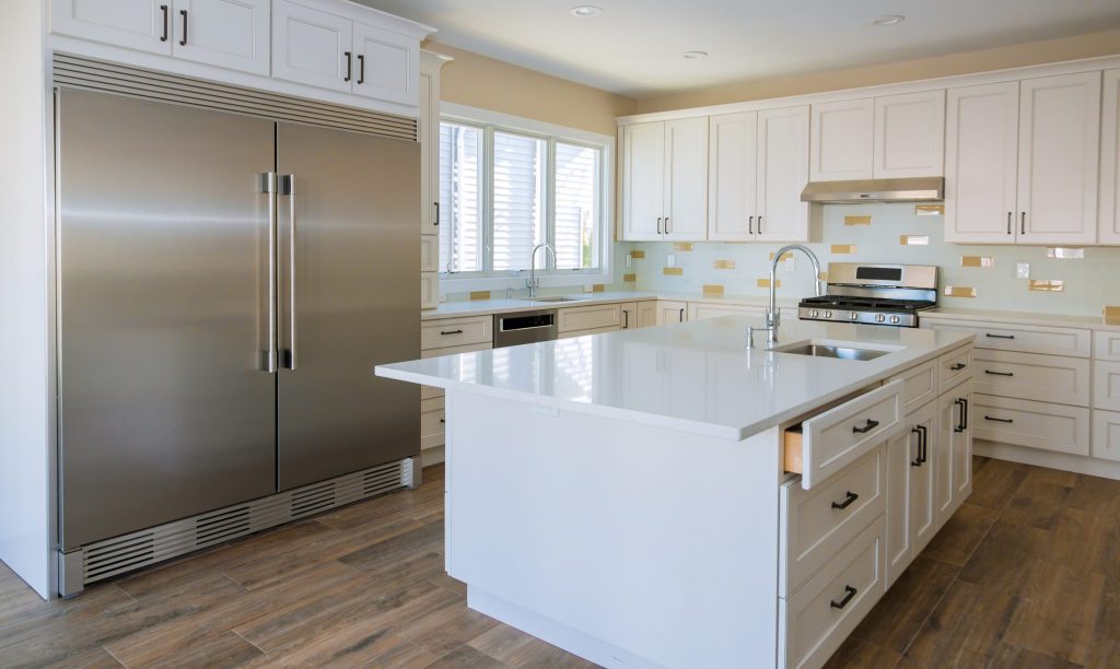 Tall stainless built-in refrigerator set flush with white cabinetry in a bright kitchen with a stone island