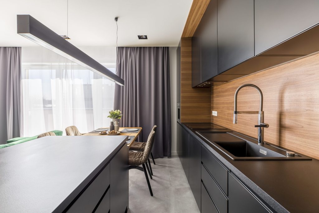 A black handleless galley kitchen with a timber splashback and a linear pendant over the island