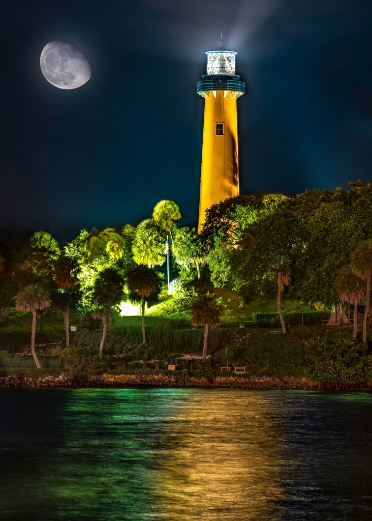 The Jupiter Inlet lighthouse lit at night above the water, with the moon behind it