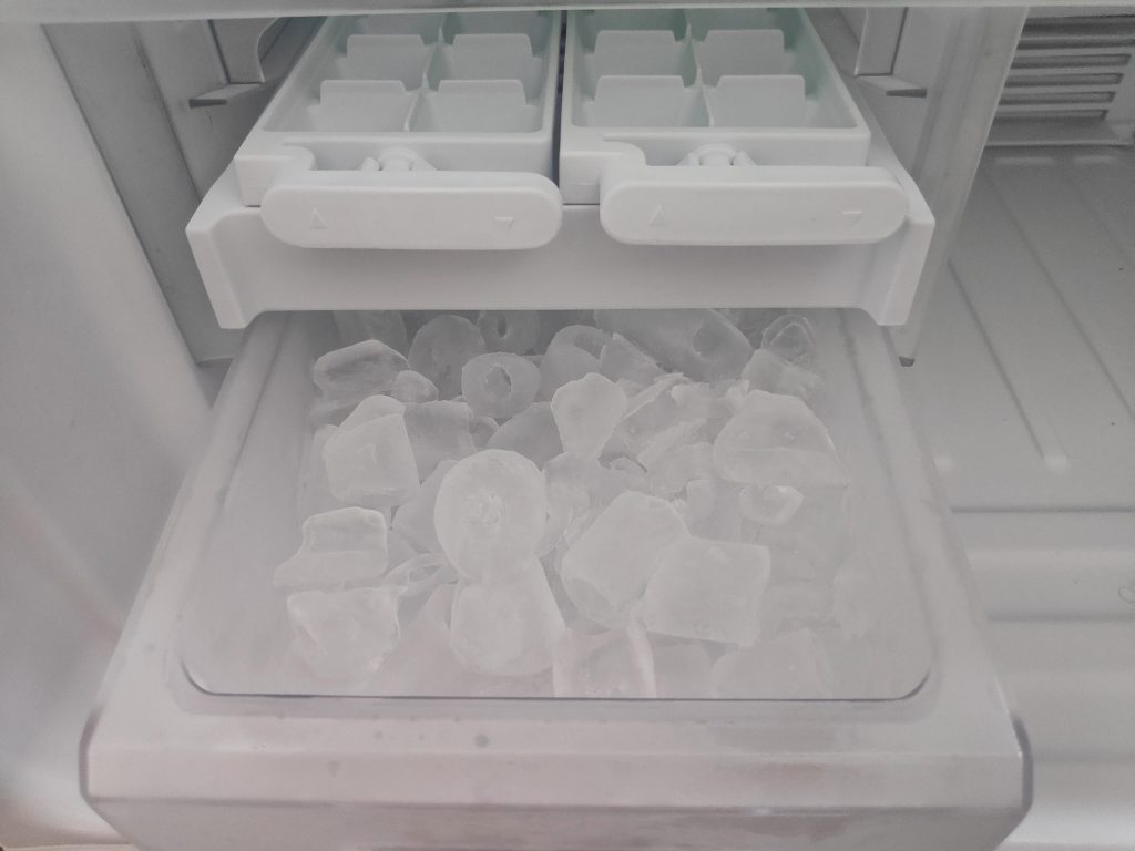 An ice maker inside a freezer, its twist trays above a bin filled with cubes