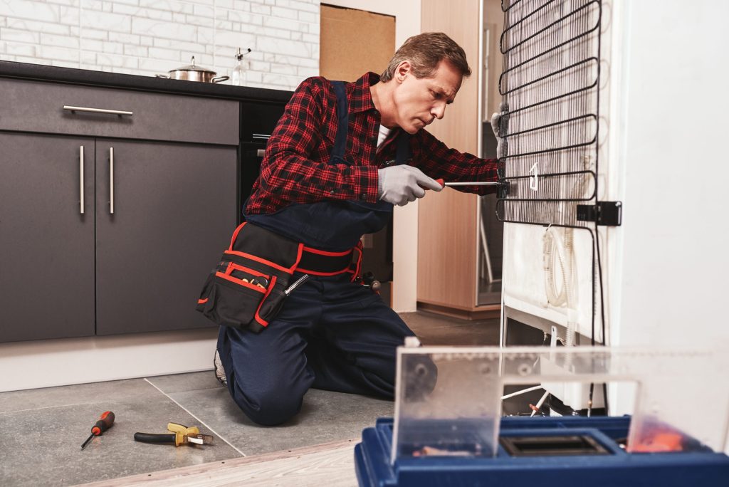 A technician working on the condenser coil at the back of a refrigerator with a screwdriver