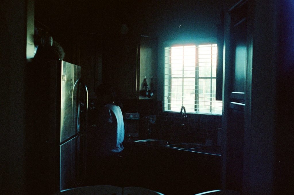 A darkened kitchen lit only by daylight through the blinds, the refrigerator standing in shadow
