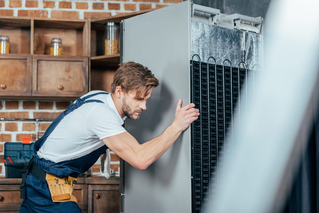A technician easing a refrigerator away from the wall, its condenser coil exposed down the back